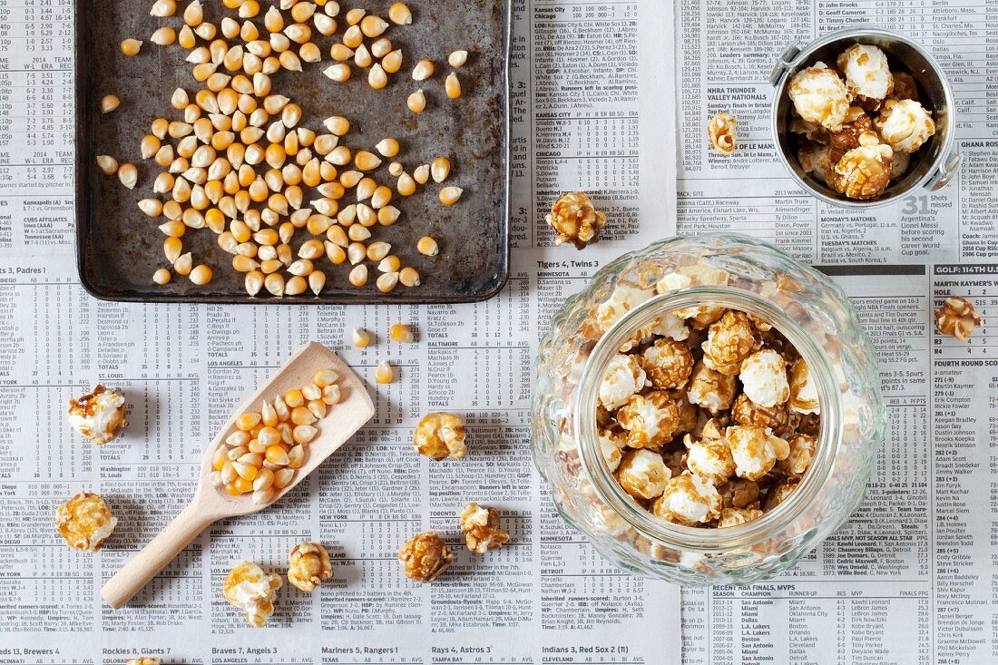 A jar of caramel popcorn next to kernels on a baking tray