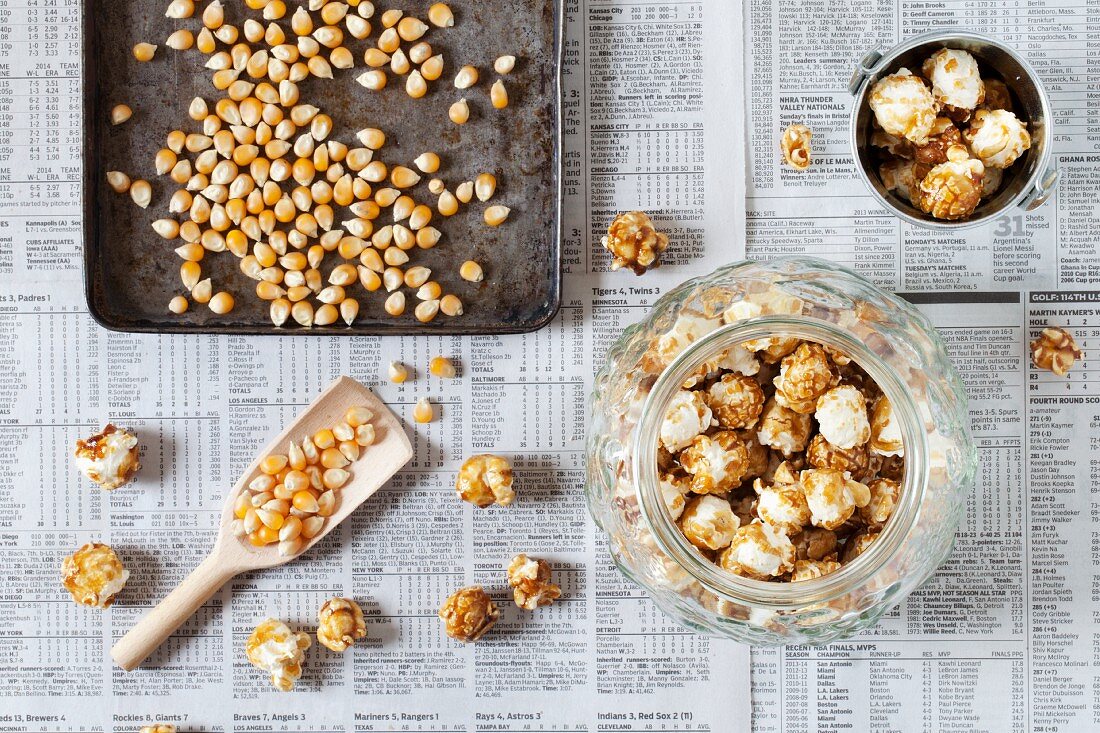 Ein Glas karamellisiertes Popcorn neben Backblech mit Maiskörnern