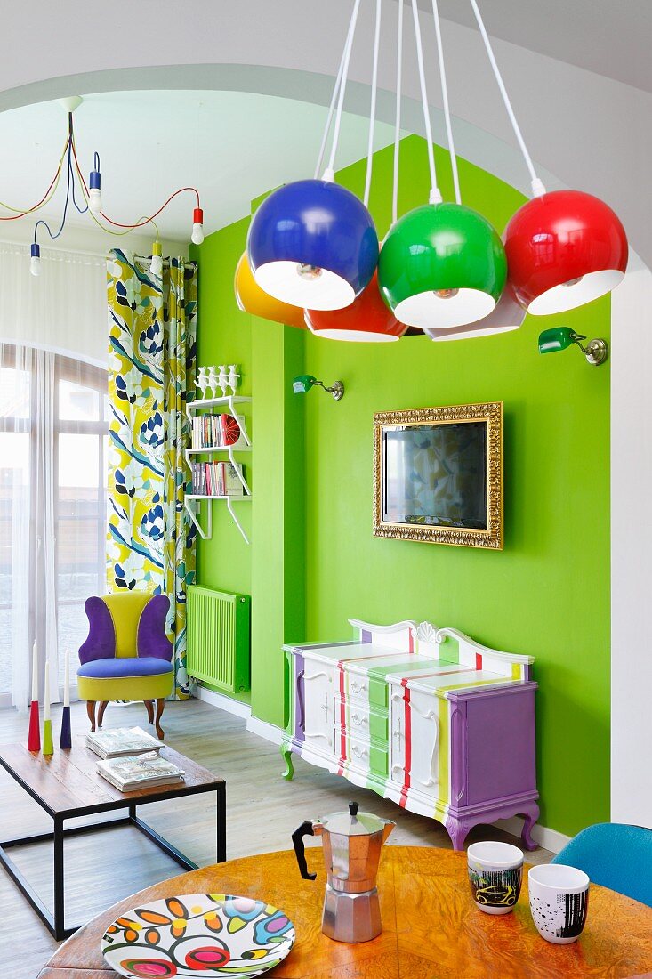 Farbenfroher Wohnbereich mit neongrüner Wand, bemalter antiker Kommode und bunten Kugelleuchten