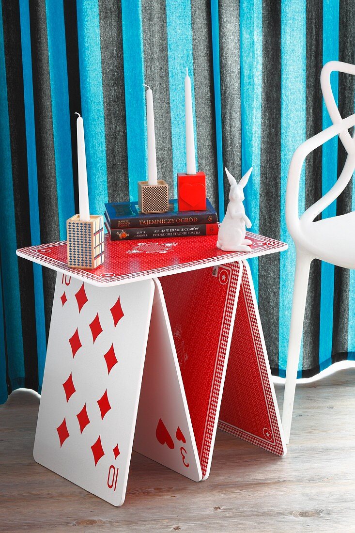 Whimsical side table in red and white with playing card motif, various candlesticks and white rabbit ornament in front of striped curtains