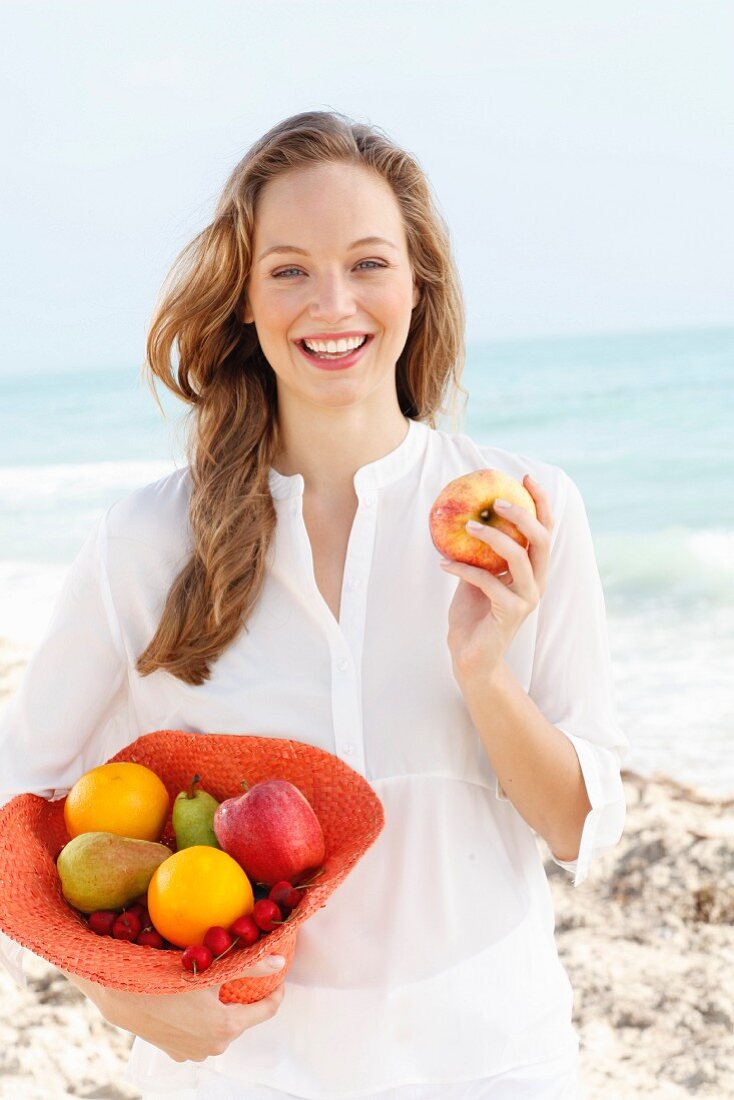 A young woman by the sea wearing a white blouse holding a hat filled with fruit