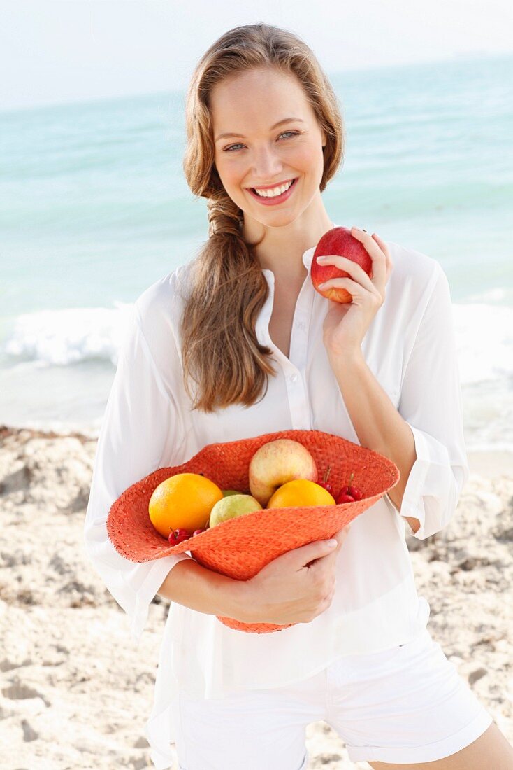 A young woman by the sea wearing a white blouse and shorts holding a hat filled with fruit