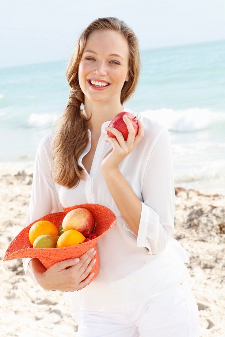 A young woman by the sea wearing a white blouse holding a hat filled with fruit