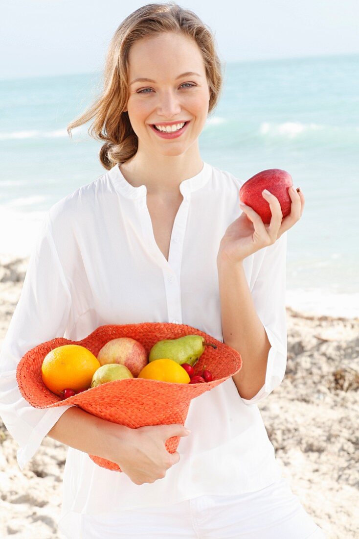 A young woman by the sea wearing a white blouse holding a hat filled with fruit