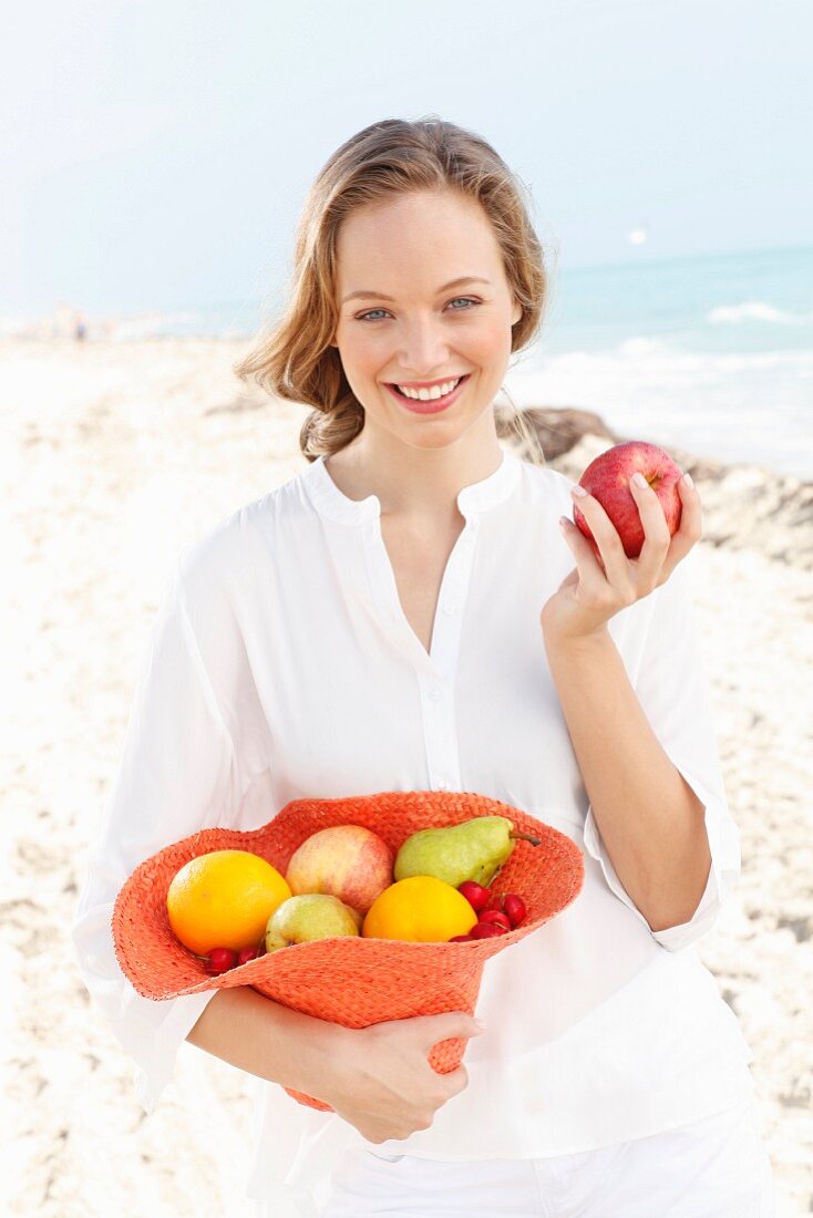Junge Frau in weisser Bluse hält mit Obst gefüllten Hut am Meer
