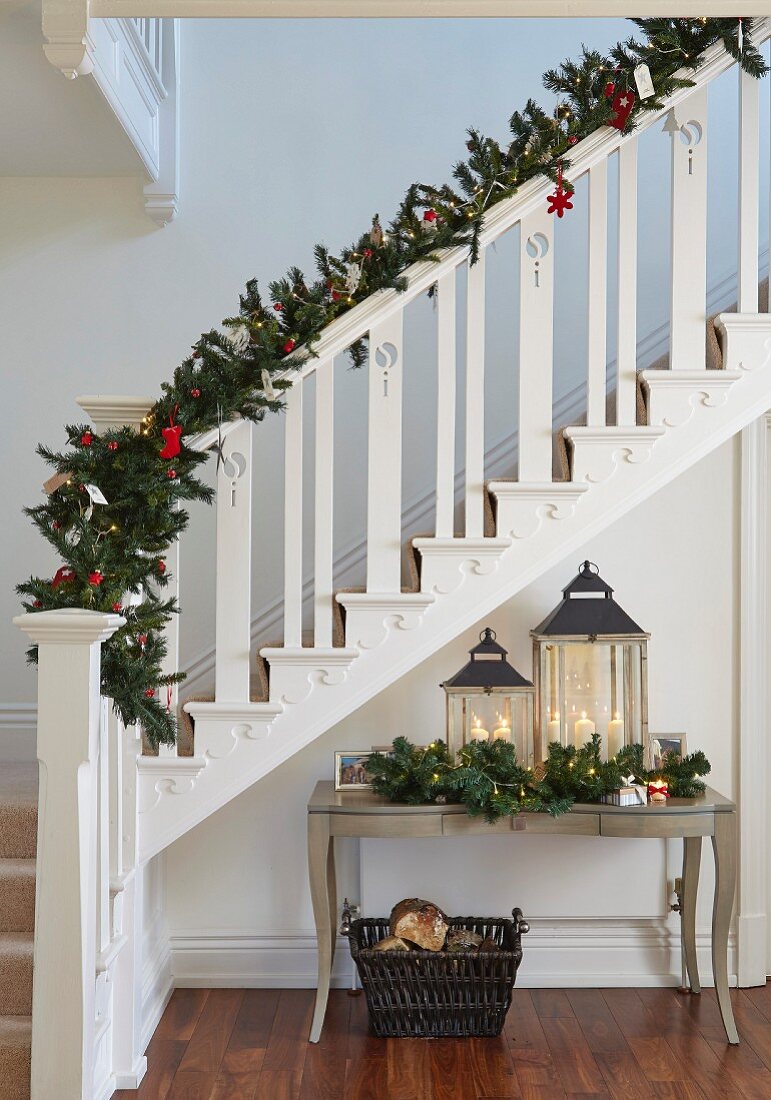 White-painted wooden staircase decorated with fir branches and Christmas decorations above lanterns on console table with curved legs