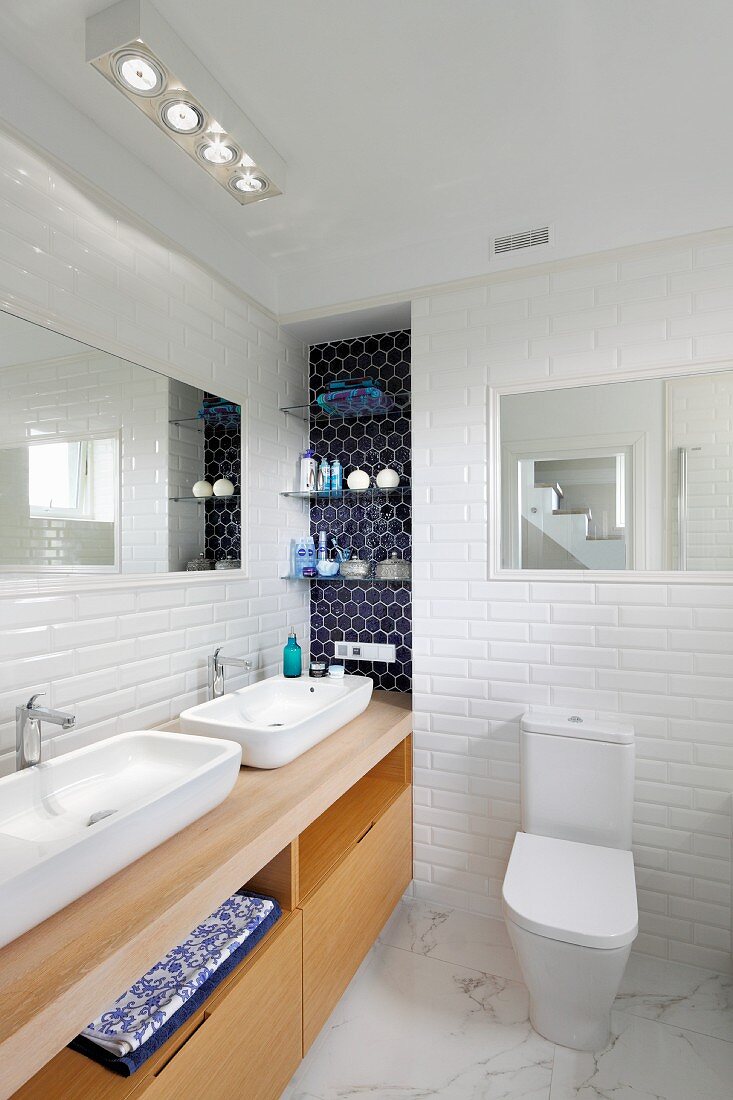 Long washstand with twin sinks on wooden base unit in white-tiled bathroom with marble floor