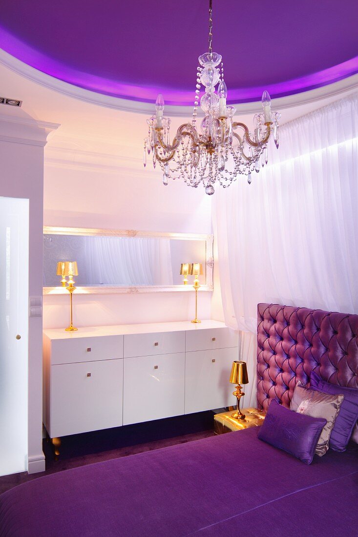 Elegant bedroom in shades of purple with gilt table lamps on white dressing table, purple ceiling panel and crystal chandelier