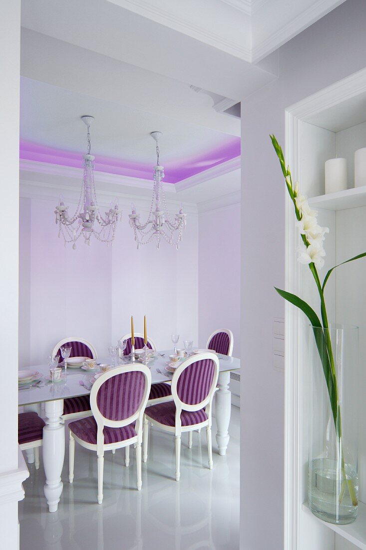 Dining table and crystal chandelier in dining room with purple, indirect lighting on ceiling and Neo-Baroque, upholstered chairs