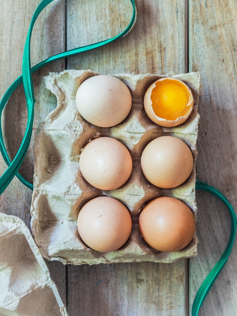 Six fresh organic eggs in an egg box, one cracked open