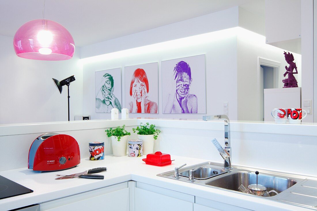 View across white kitchen counter to photo portraits on white wall and pink pendant lamp