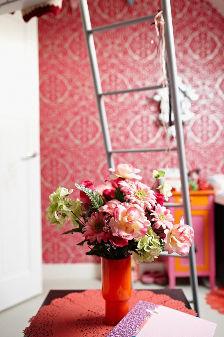 Bouquet in orange glass vase on doily in front of metal ladder and pink, patterned wallpaper