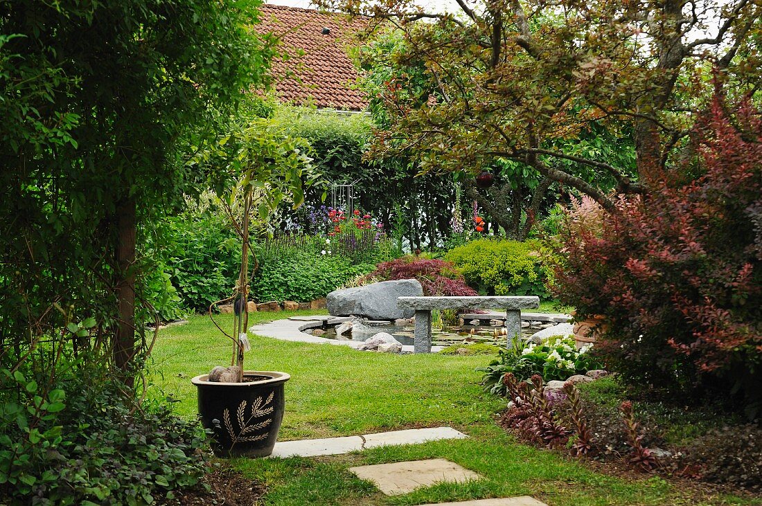 Summery garden with small potted tree, stone bench and boulders beside round garden pond