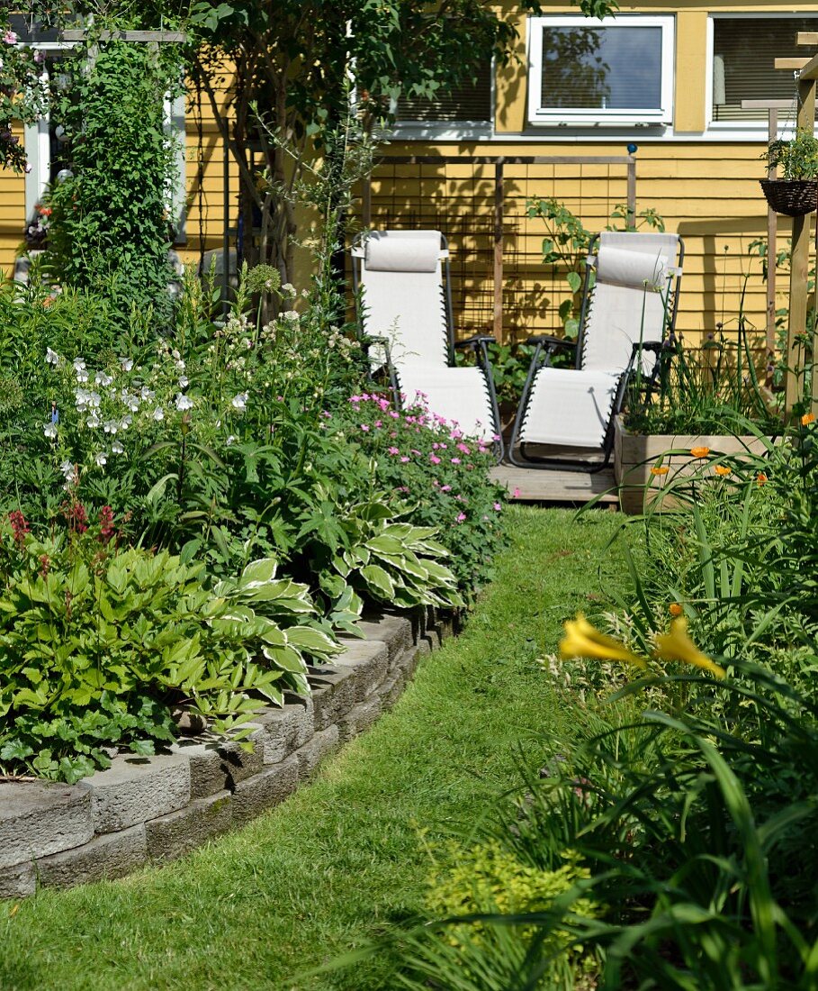 Hochbeet mit Steineinfassung, im Hintergrund Sonnenliegen vor gelb gestrichenem Haus, in sommerlichem Garten