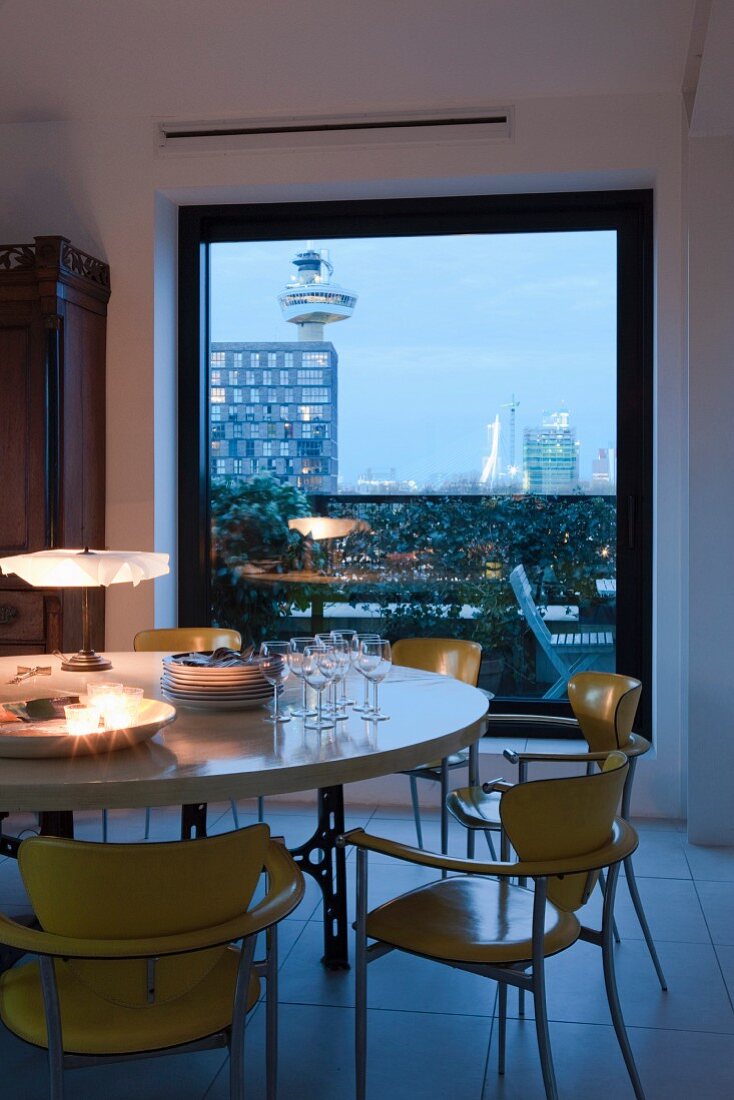 Romantically lit dining table with retro chairs in front of floor-to-ceiling window with view of city at twilight