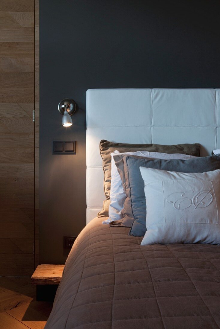 Multiple scatter cushions on bed with headboard upholstered in white against dark grey bedroom wall with sconce lamp