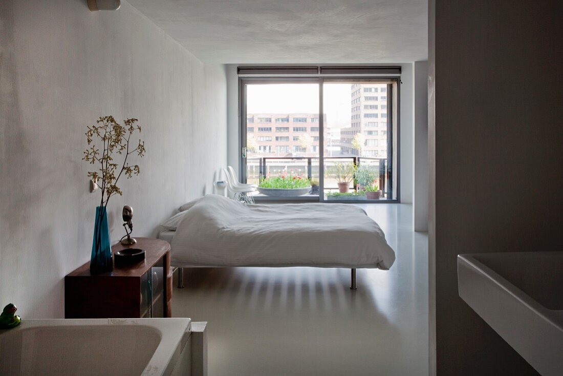 View from ensuite bathroom into minimalist bedroom with double bed and French windows leading to balcony