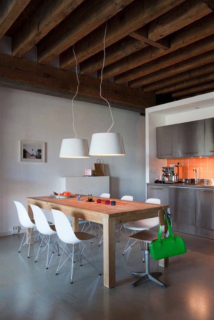 Edelstahl Küchenzeile mit orangefarbenen Wandfliesen, rustikale Holzbalkendecke und Holztisch mit weißen Designerstühlen in Loftwohnung