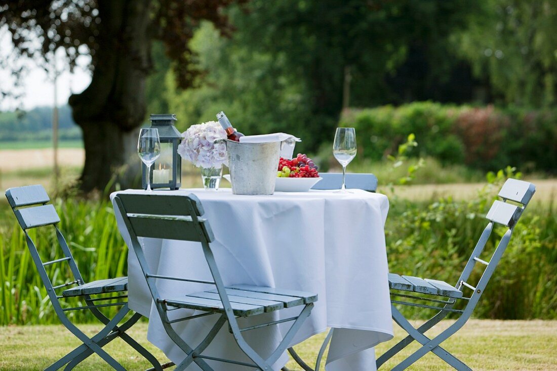 A garden table laid with a white table cloth, a wine cooler, wine glasses, a hydrangea and a fruit bowl