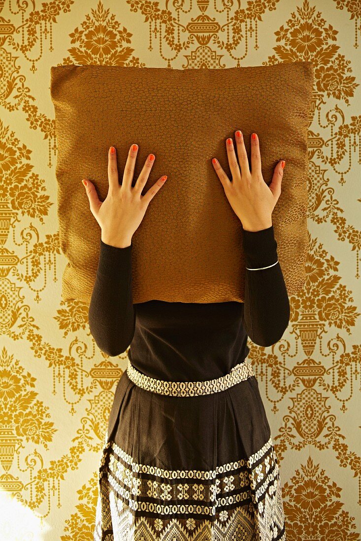 Woman with painted fingernails holding cushion in front of face stood against wallpaper with gold, ornate pattern