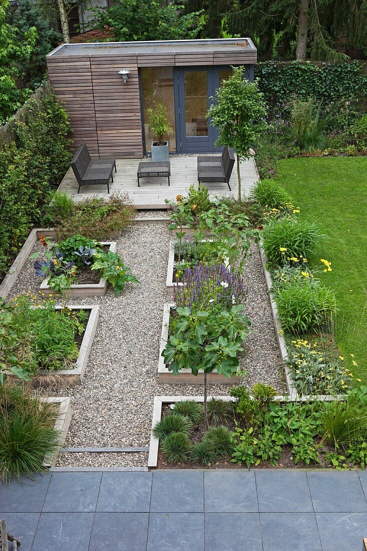 Raised beds in geometric arrangement with gravel paths in front of modern outdoor furniture on terrace and summer house