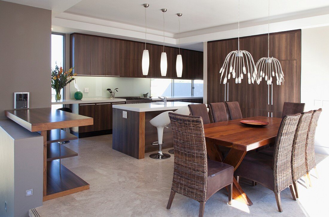 Dining area with solid wooden table and wicker chairs with seat cushions in front of open-plan fitted kitchen with exotic wood fronts and retro lamps