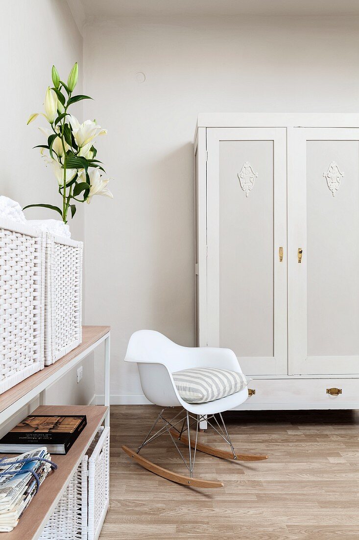 White, designer rocking chair next to painted wardrobe, bouquet of lilies and white storage baskets on open-fronted shelving in corner of bedroom