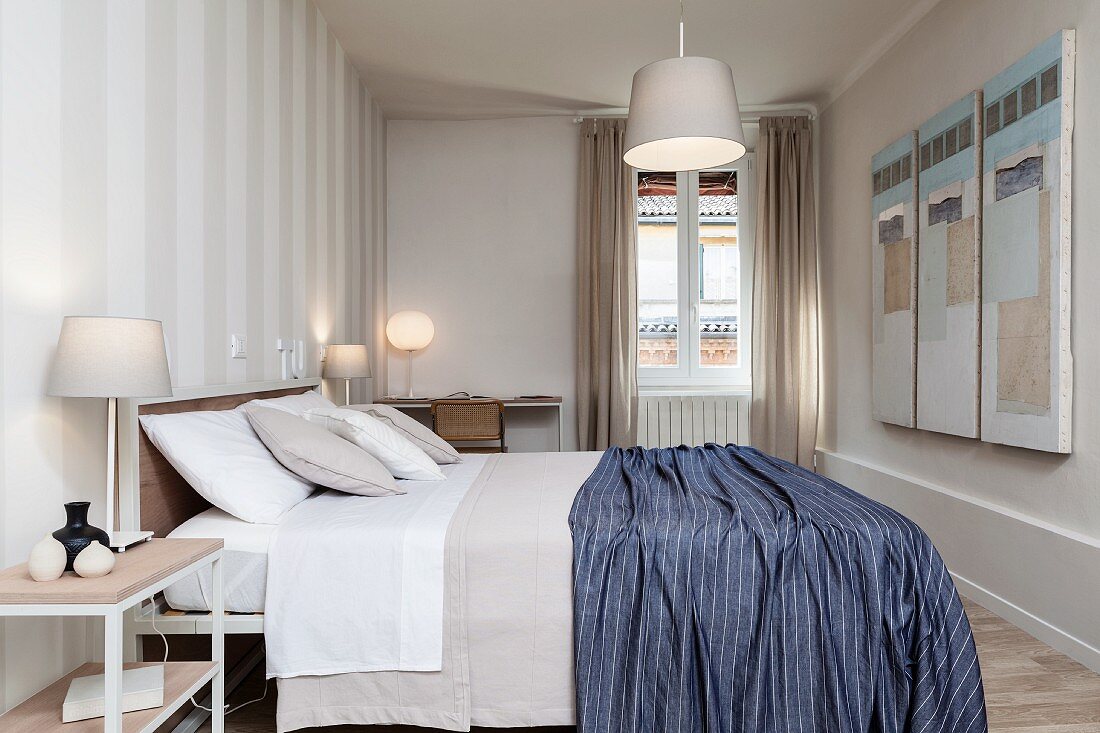 Bedroom with white and grey striped wallpaper, blue blanket on double bed, desk and various lamps