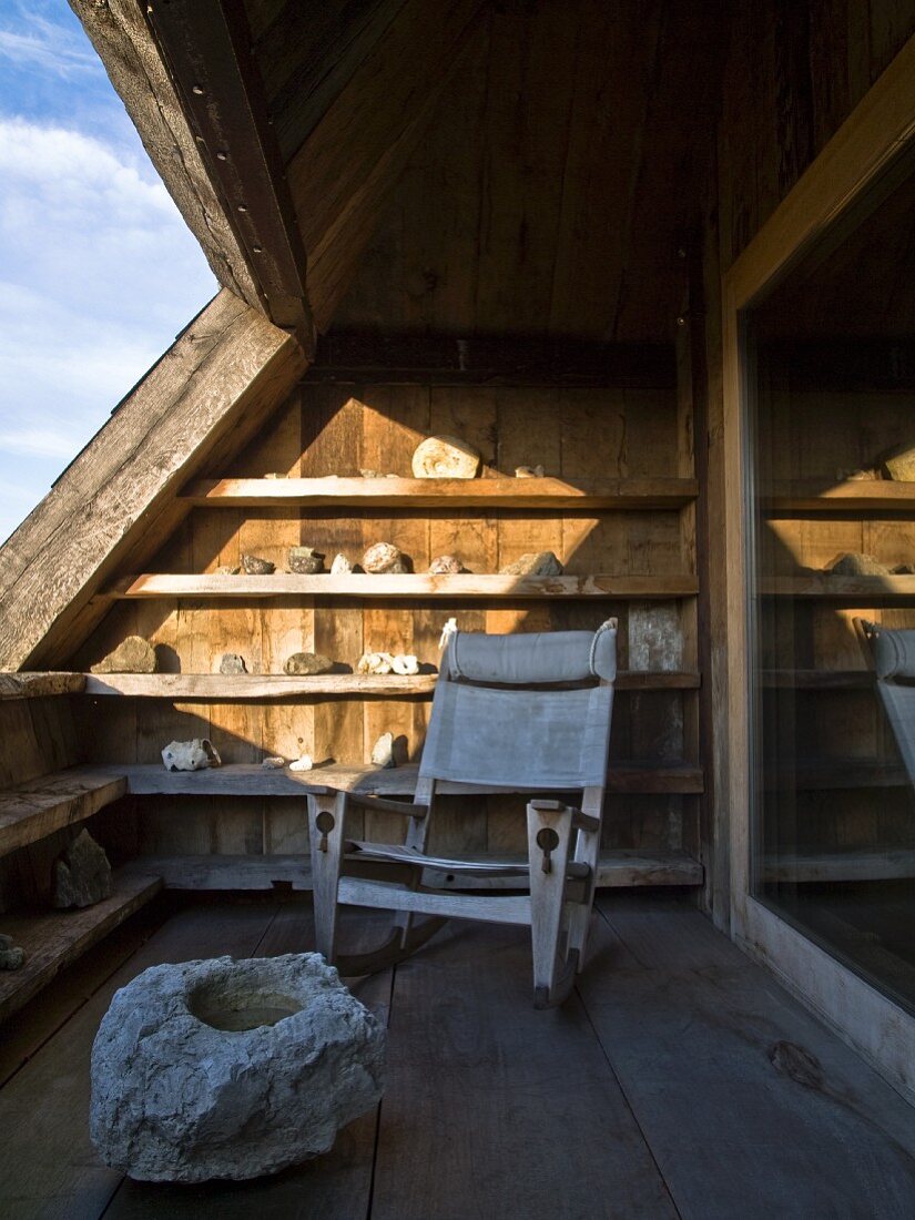 Rustic armchair with canvas cover on roof terrace below shelves of rocks in patch of sunlight and boulder in foreground on wooden floor
