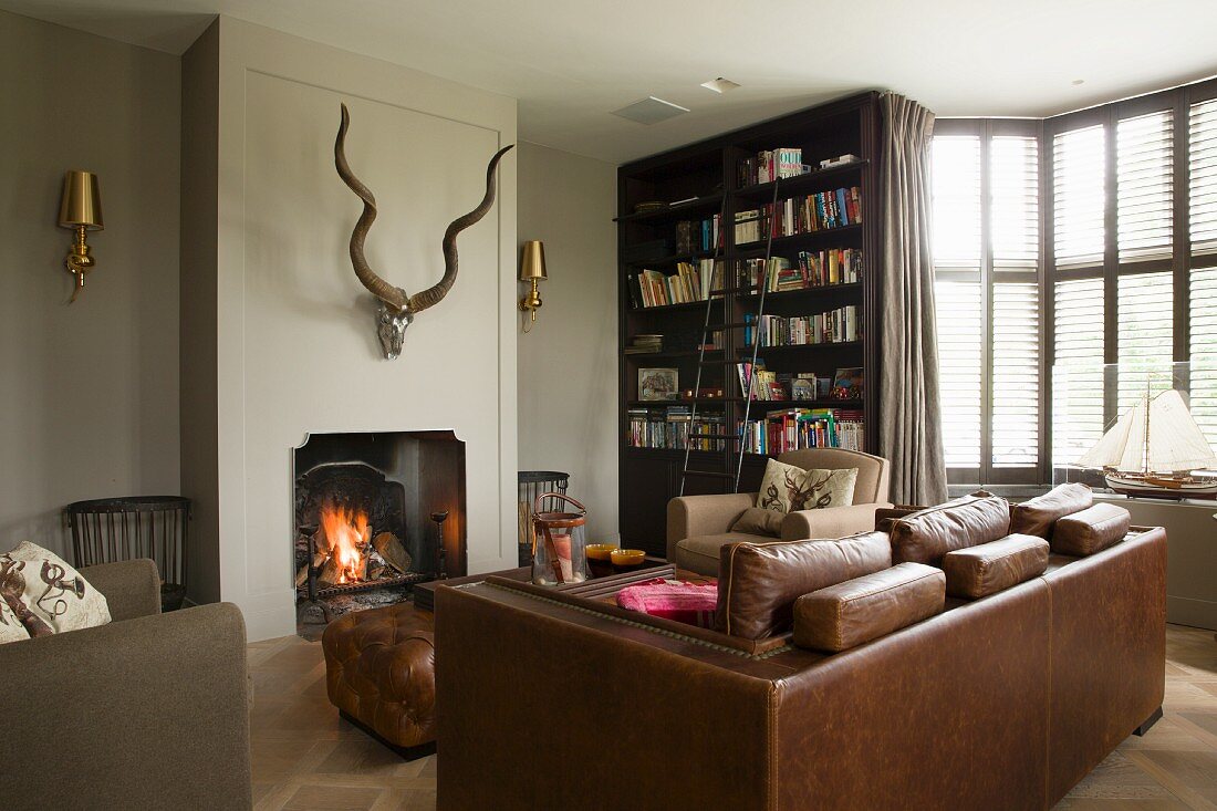 Brown leather sofa and armchair in front of fire in open fireplace below hunting trophy in elegant interior