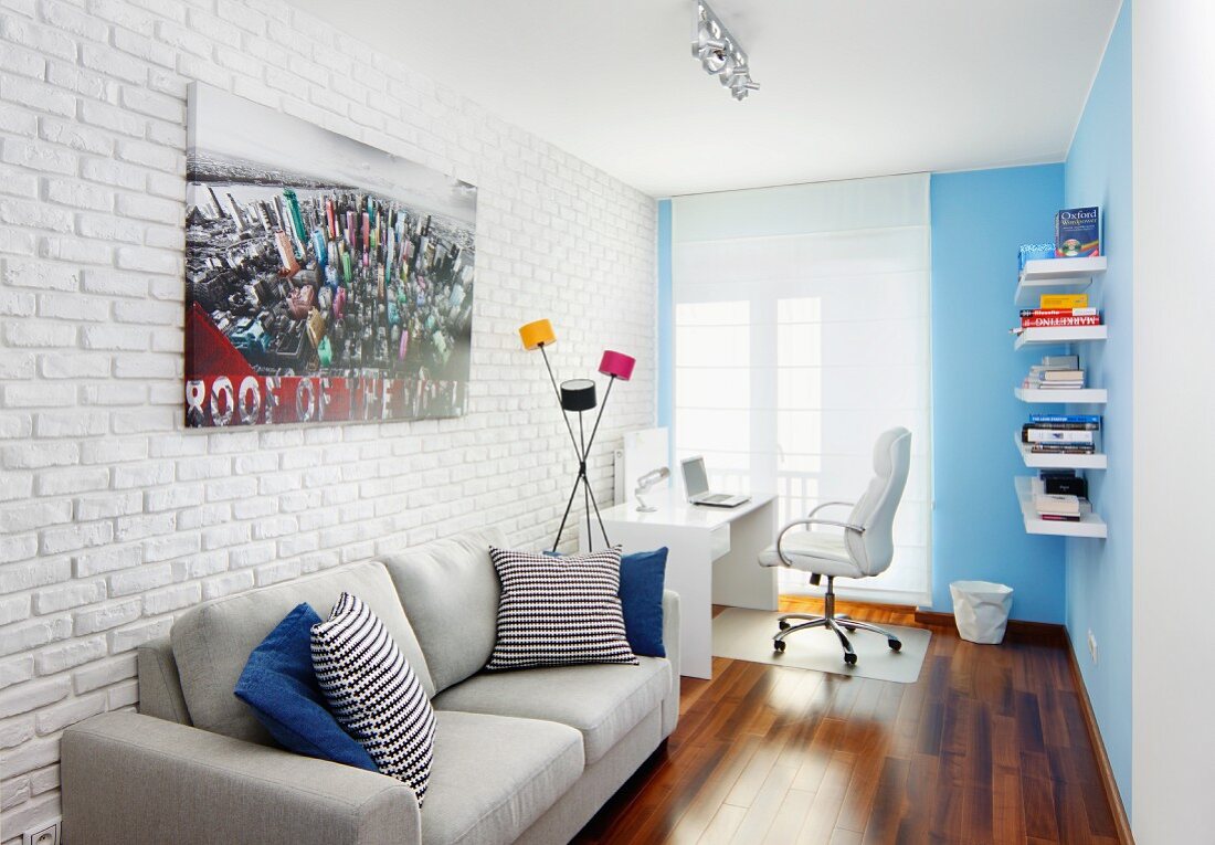 Pale grey sofa with scatter cushions against whitewashed brick wall, retro standard lamp next to desk and white office chair