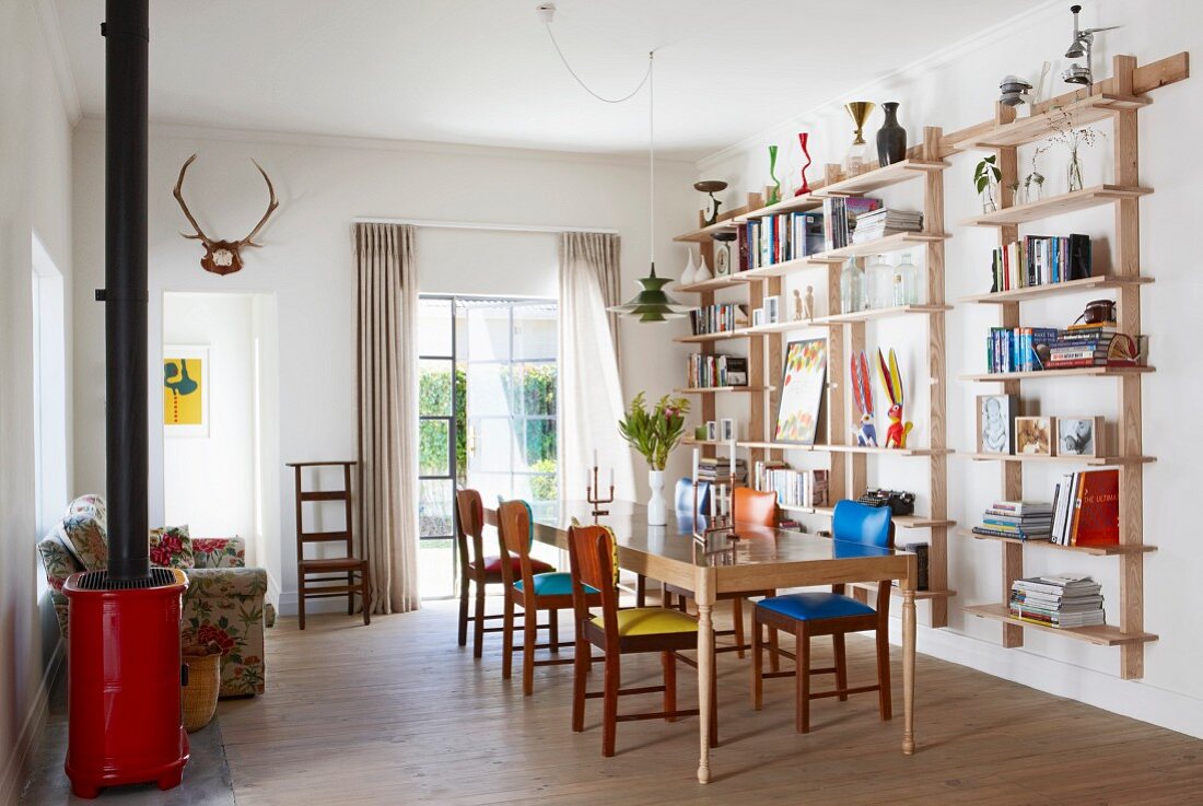 Books on light shelving, retro chairs with different coloured seat cushions around dining table, red log burner and floral sofa