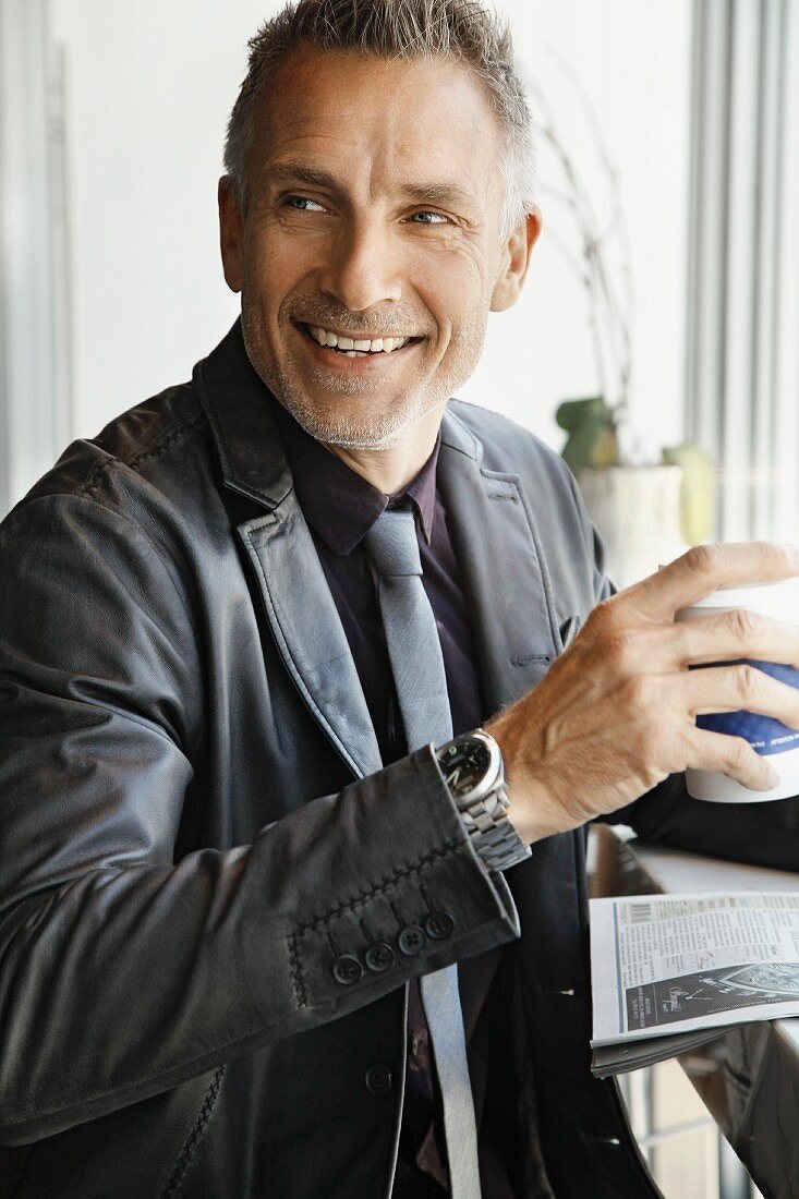 A middle-aged man wearing a black shirt, a tie and a leather jacket with coffee and newspaper