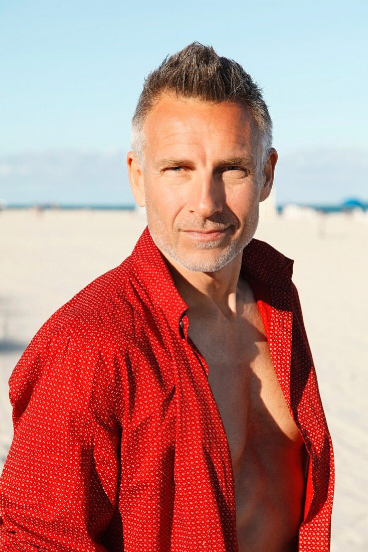 A middle-aged man on a beach wearing a red, unbuttoned shirt