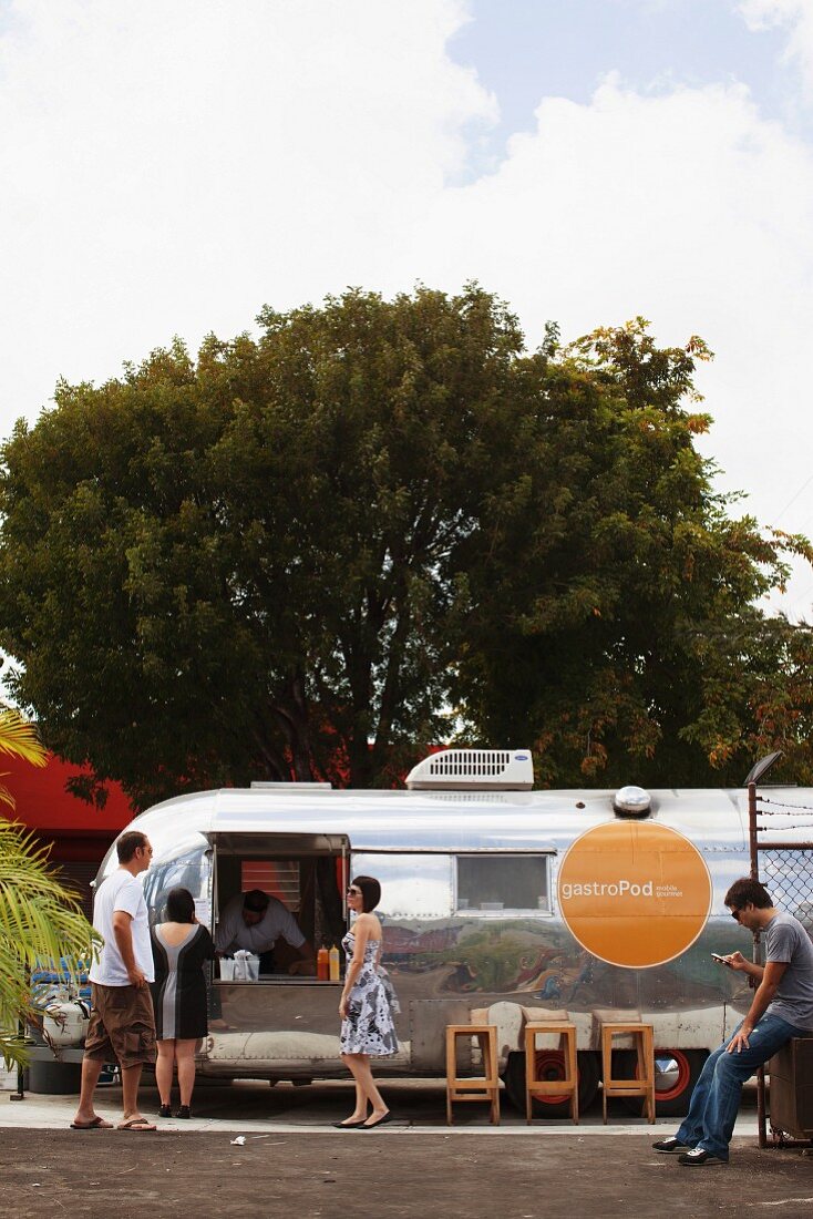 A shiny silver fast food car with people waiting to be served