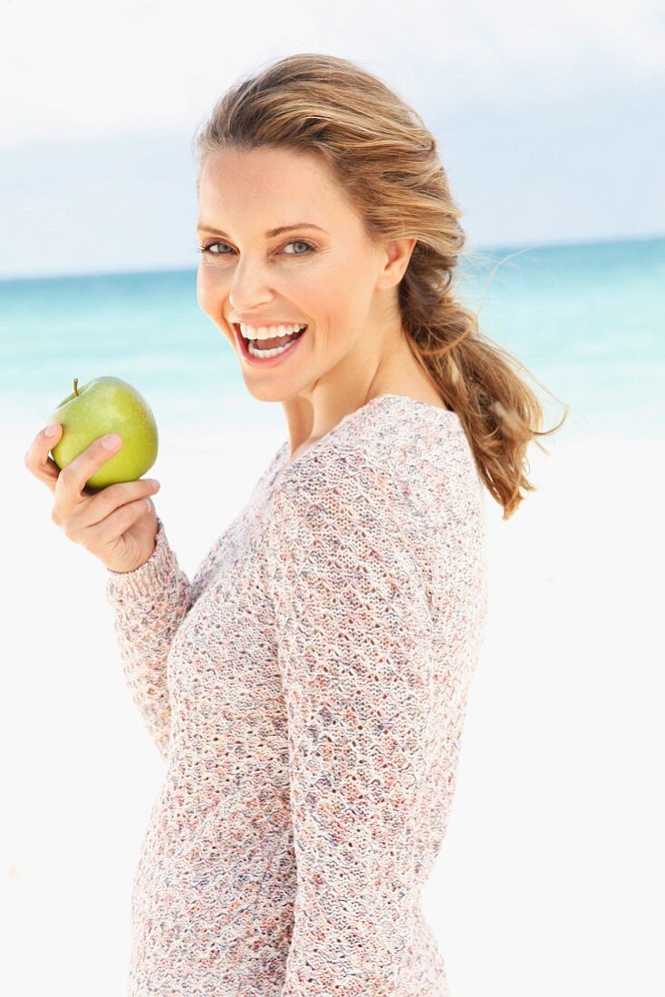 Blonde Frau in gestricktem Longpulli hält einen Apfel in der Hand am Strand