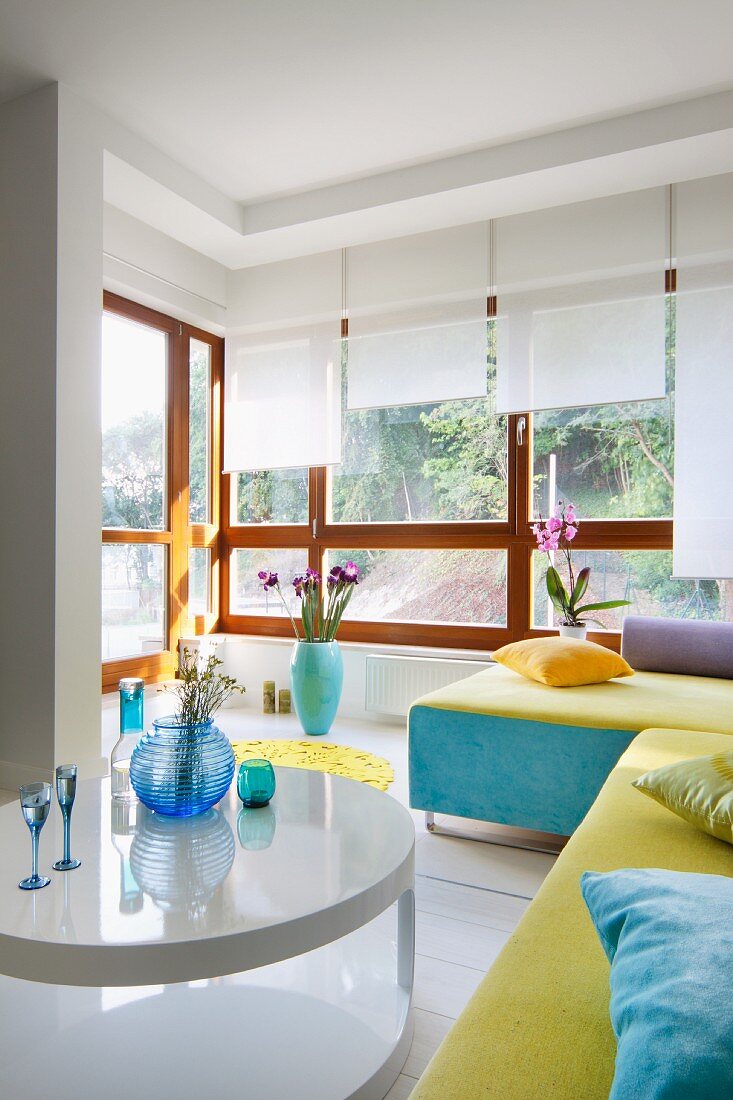 White coffee table in front of couch with blue and yellow cover in front of bay window with wooden frame and white roller blinds