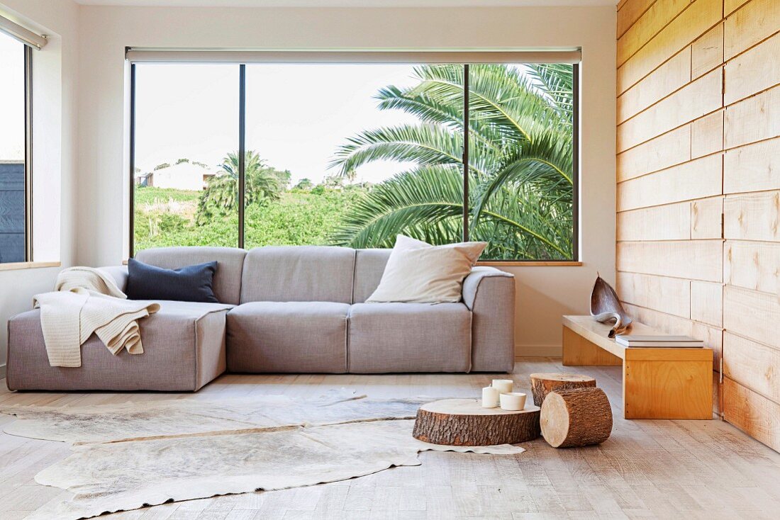 Comfortable seating area with sofa next to panoramic window, animal-skin rug on wooden floor, slices of tree trunk used as low coffee tables and bench against wood-clad wall
