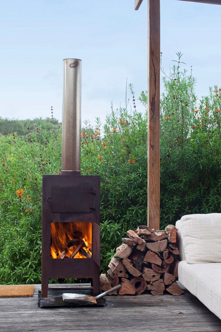 Kaminofen mit Feuer neben kleinem Brennholzstapel auf Terrasse, seitlich teilweise sichtbare helle Couch, im Hintergrund dichtbewachsener Garten