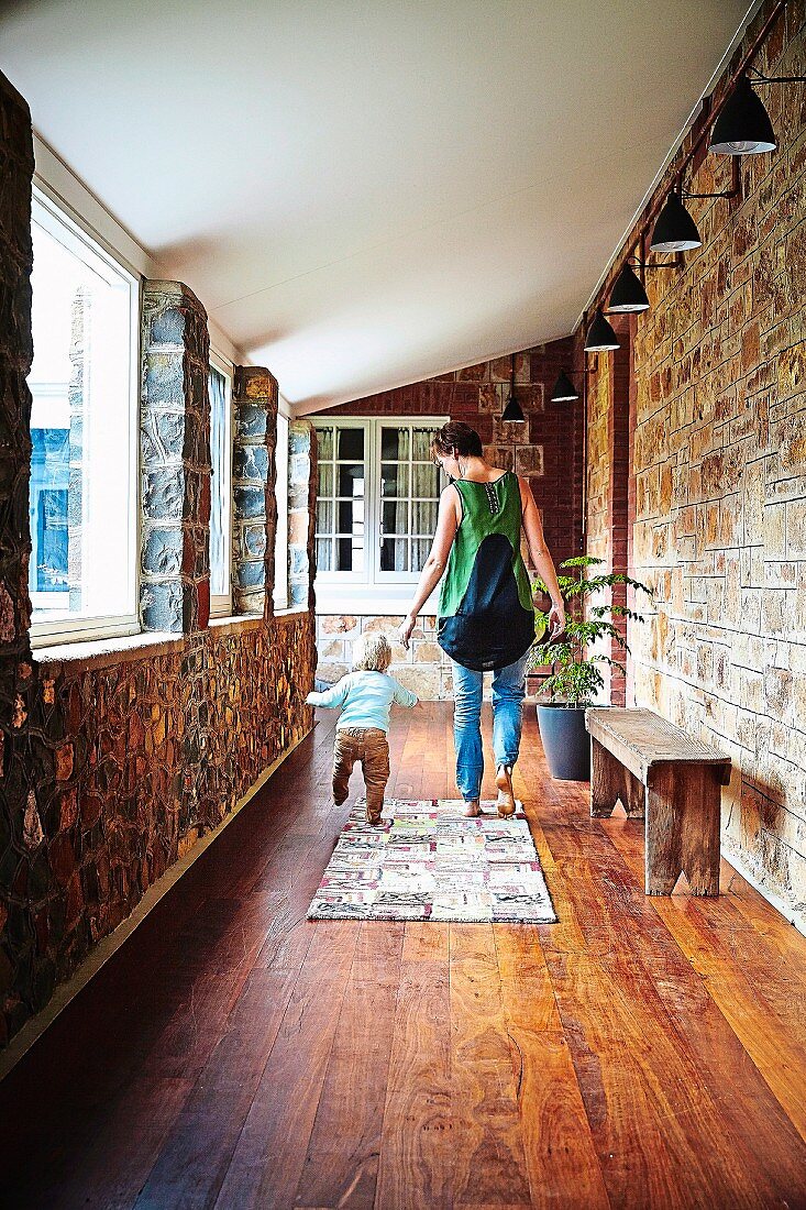 Woman and toddler walking along veranda with wooden floor and wooden bench below row of wall-mounted lamps
