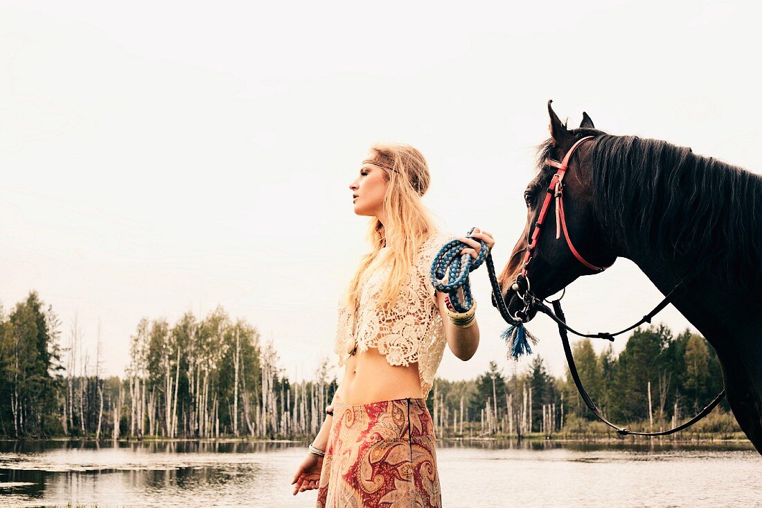 A young blonde woman wearing hippie-style clothes leading a horse by a lake