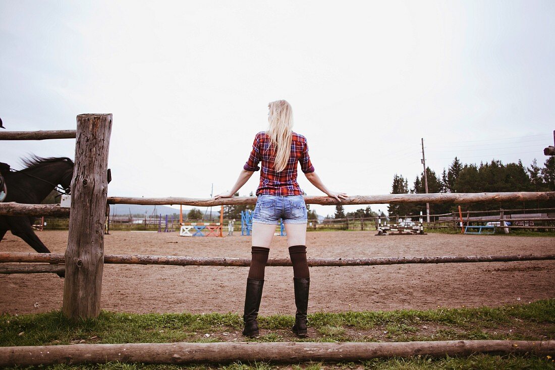 A long-haired, blonde woman wearing a checked shirt and short shorts standing on a wooden fence