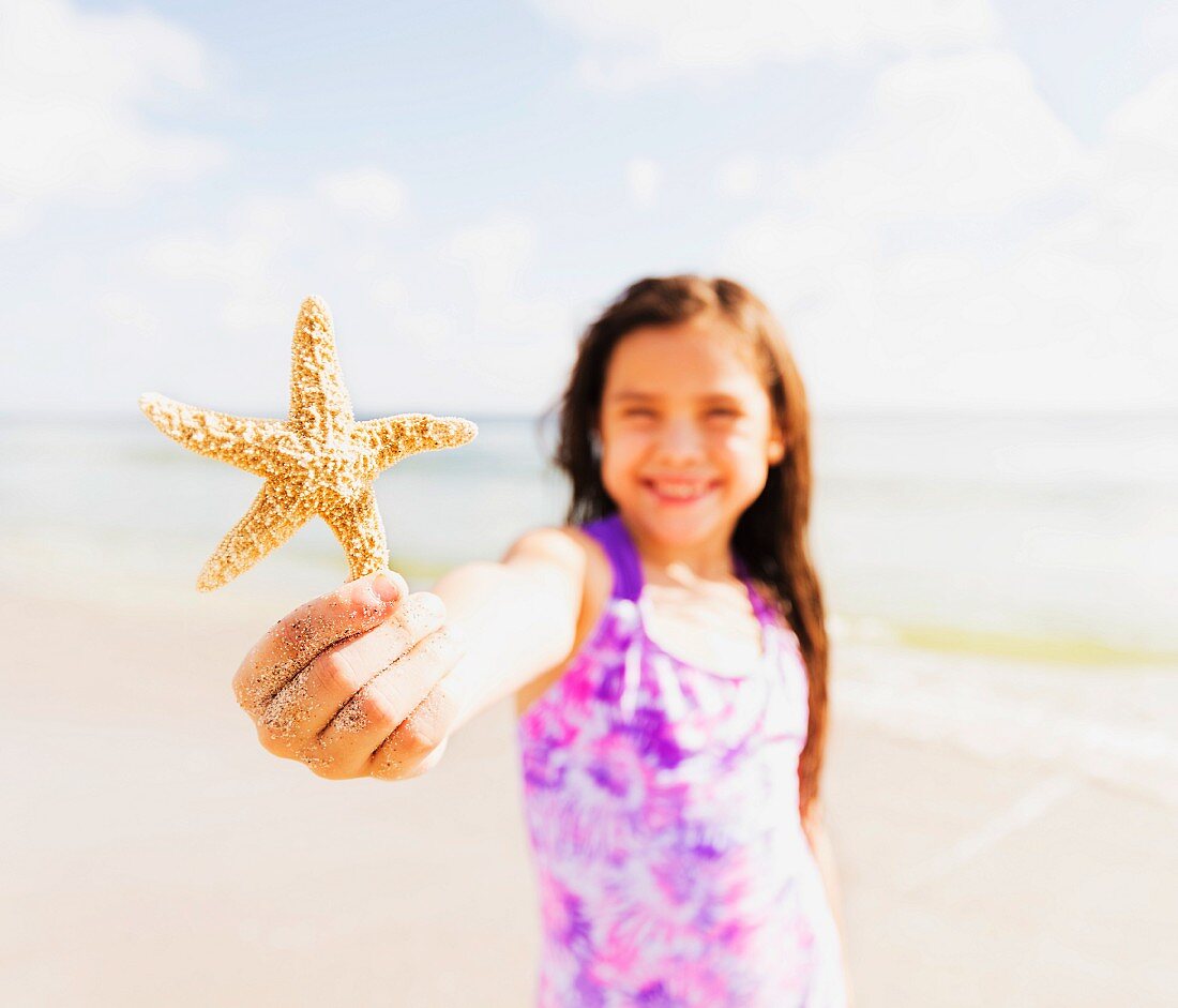 A girl on a beach wearing a purple patterned dress holding a sea star