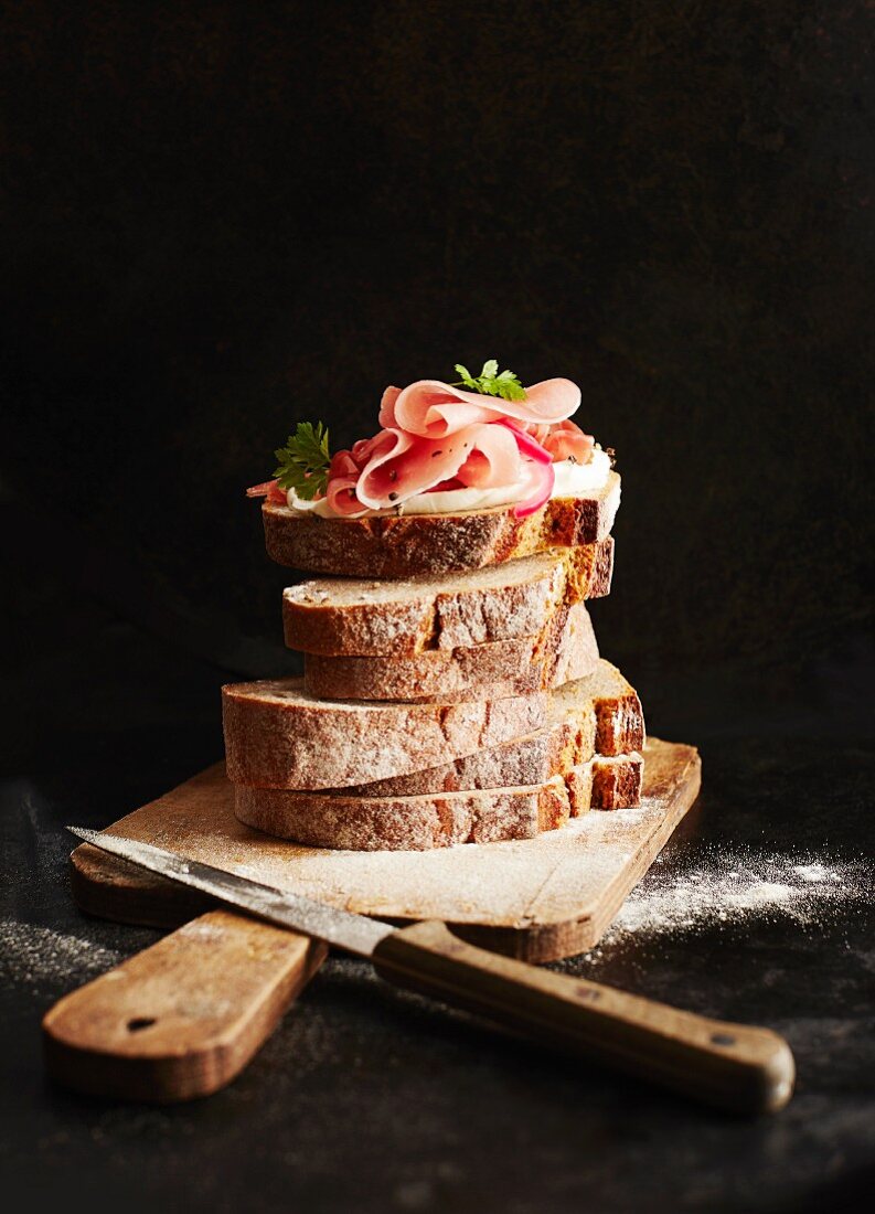 Brotscheiben auf einem Holzschneidebrett; obenauf ein Schinkensandwich