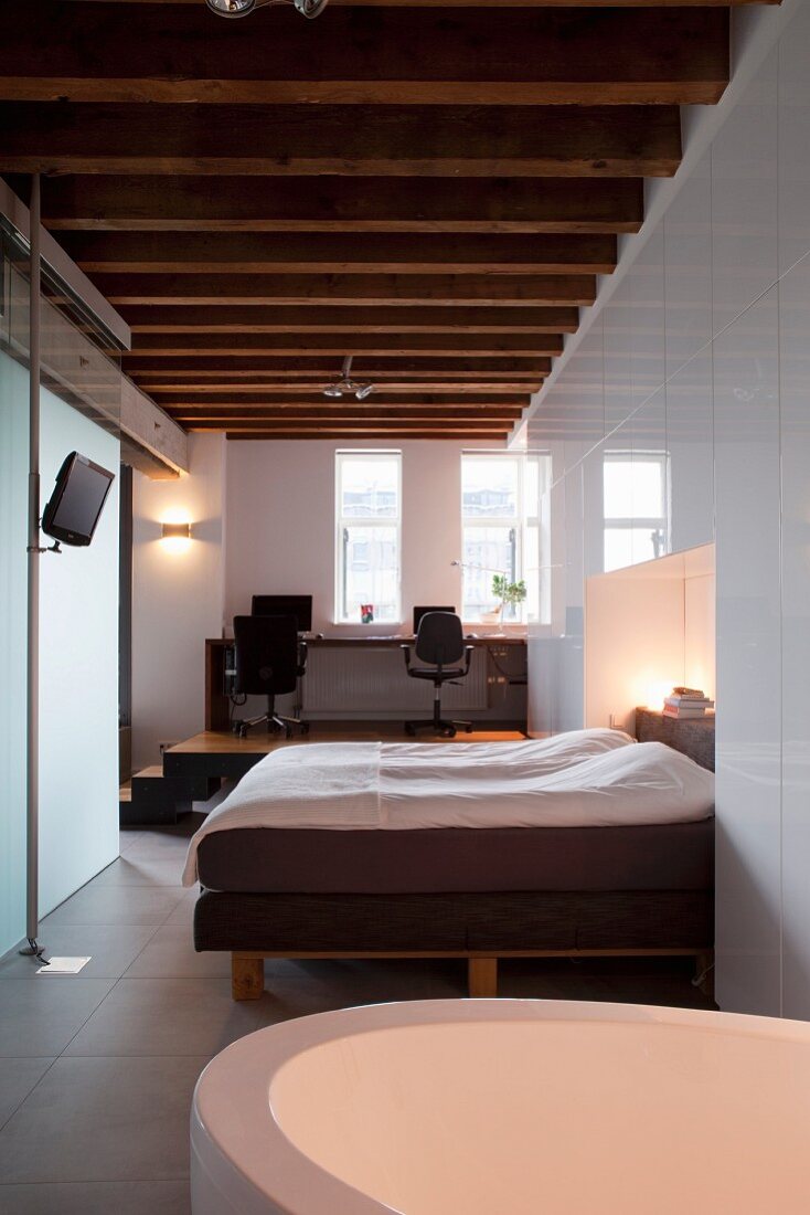 Bathtub in bedroom and double bed in niche formed by white fitted wardrobes; view of office on platform in elegant, renovated loft apartment