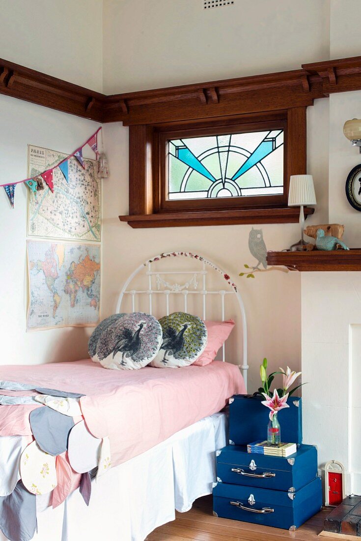 Blue suitcases stacked on floor and used as bedside table next to white metal bed in corner below transom window with Art-Nouveau motif