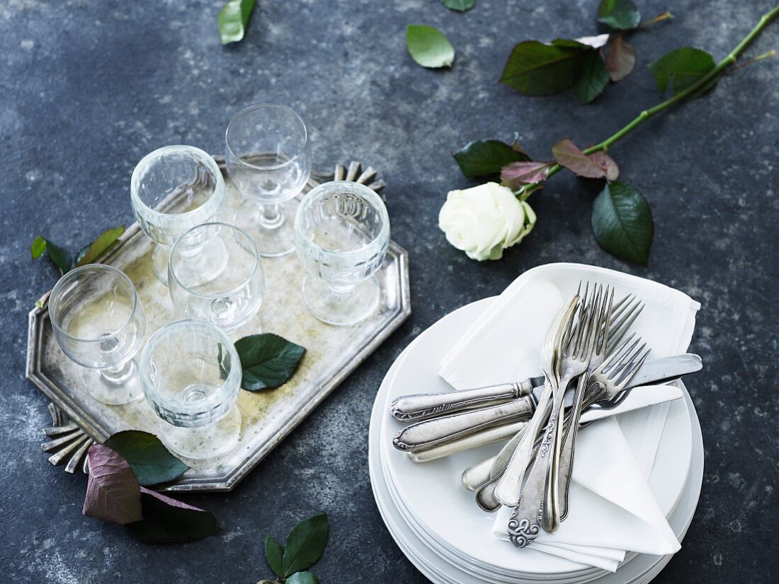 A stack of plates with napkins and cutlery next to glasses and a rose on a tray