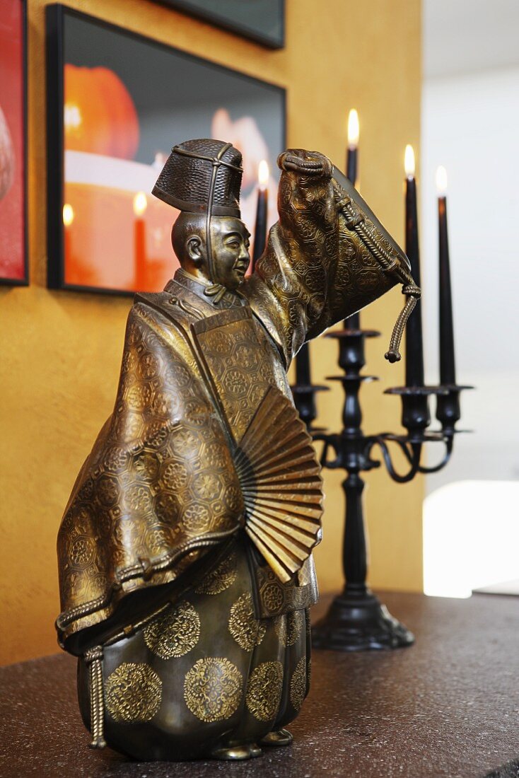 Oriental, metal figurine and lit candles in candelabra on surface below framed pictures on yellow wall