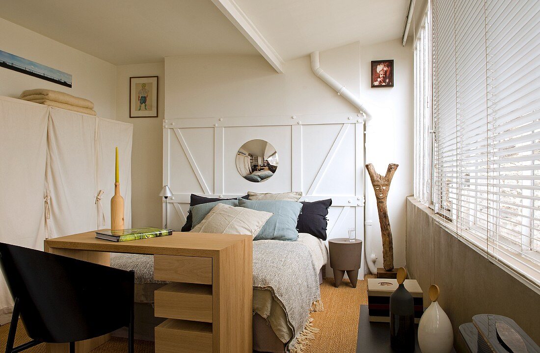 Desk at foot of bed against white, wooden wall in rustic, modern bedroom with closed louver blinds on windows