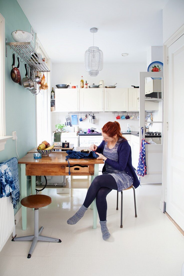 Junge Frau am Küchentisch vor Wand mit Regalbord und aufgehängtem Kochgeschirr, im Hintergrund weiße Einbauküche